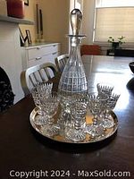 Full view of the tear-shaped crystal decanter with stopper surrounded by seven matching cordial glasses on a round metal tray, displayed on table.