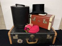 Group photo of all vintage luggage and hats, showing black McBrine luggage at base, cylindrical hat box, Constellation makeup case, black top hat, and two floral women's hats.