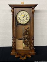 Full front view of the wooden vintage Rensie pendulum wall clock showing turned columns, glass door with pendulum, and detached top piece.