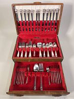 Full view of open chest with all flatware pieces organized inside on red velvet lining showing knives, forks, spoons with floral handle detail.