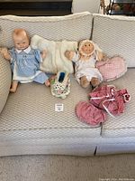 Two dolls sitting on a beige couch with a white mink coat behind them, a crocheted slipper in front, and pink doll clothing laid out beside them.