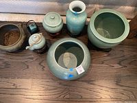 Full set of green ceramic pots and vases with one brown decorated planter, placed on wooden floor.