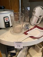Photo showing rice cooker, glass measuring cup, hand mixer with beaters, cheese grater, plastic spatula, and manual sharpener on a white round table.