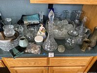 Overview of the assorted kitchen glassware and decor showing large crystal decanter, punch bowl with cups, various vases, a ceramic figurine and small decorative items on a countertop.