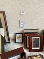 Several picture frames grouped on carpet near wall outlet. Frames include wood and black finishes in various sizes stacked against wall.