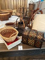 Photo showing multiple wicker and woven baskets, focus on decorative chest-style basket and various oval and round baskets with handles.