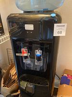 Front view of black Aquverse water cooler with red and blue taps and drip tray, with 5-gallon water bottle inverted on top. Located in kitchen pantry.