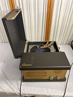 Front angle showing turntable open with visible record and tonearm inside, unit closed front panel with speaker grill and knobs visible.