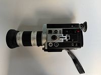 Side profile of vintage Canon Super 8 film camera showing long zoom lens and textured black/metal body with wrist strap.