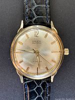 Close-up of the watch face showing Gruen Precision dial, gold tone hands and markers, black faux crocodile leather strap with blue stitching.