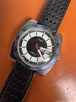 Front view of the watch showing the dial, day and date window, red second hand, and world timer bezel with city names.