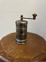 Front view of the cylindrical pepper mill grinder with silverplate body and wooden handle crank on top, placed on a wood surface.