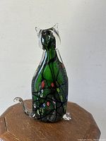 Front view of the cat figurine showing emerald green and black swirl body with clear glass ears and tail, colorful flower millefiori patterns inside.