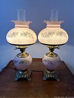 Pair of illuminated pink hued Gone With The Wind style glass table lamps with floral decorations, showing warm light diffusion, on wooden surface.