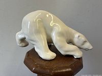 Glossy white ceramic sculpture of a polar bear on a wooden stand, angled view showing the left side of the bear.