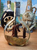 Photo showing three vintage bottles: front clear bottle contains a wooden articulated skier figurine with small carved trees and fence; two bottles behind include a dark round bottle with alarm clock and a hand painted ceramic bottle with human face design.