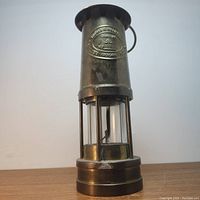 Full front view of brass miner's lamp with visible glass chamber and top vent. Maker's badge and hook visible.