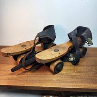 Front angle view of antique wooden roller skates showing wood bottoms, wheels, and black leather straps with buckles.