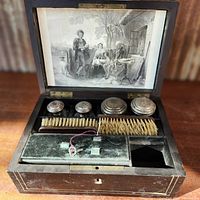 Open black lacquered travel case showing four original perfume bottles with silver lids, two brushes, and velvet compartments with a printed family scene on the inner lid.