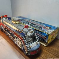 Metal toy train with colorful lithograph details and original box, captured at an angle showing front and side of the train and box.