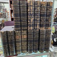 Seven faux book-style stereoview card storage containers arranged vertically on a glass shelf, showing varying wear and gold-embossed titles from 'Stereographic Library' series by Keystone View Co.
