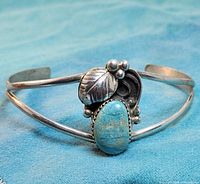 Front view of the sterling silver cuff bracelet with large turquoise stone and decorative silver leaf and bead design on a blue background.