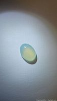 Oval cabochon chalcedony gemstone with arctic teal color placed on a white background, showing translucence and smooth polished surface.