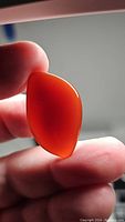 Close-up photo of an orange-red carnelian gemstone held between fingers, showing smooth polished surface, translucency, and organic fancy cabochon shape.
