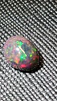 Close-up image showing the oval cabochon black opal with strong multicolored flashes against a black textured fabric background.