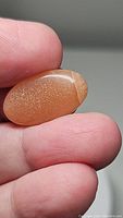 Close-up photo of the natural peach moonstone oval cabochon stone held between fingers showing its color, shape, and sparkling inclusions.