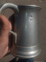 Close-up side view of aluminum tankard showing stamped markings including a small circular emblem and letters near the rim.