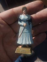 Front view of 3-inch plastic figurine depicting Laura Secord, showing details of dress and facial features, held in hand for scale.
