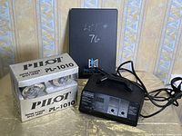 Photo showing Eliminator Intelligent Battery Charger with digital display and power cord, alongside two boxed PILOT Super Power Spot Lamps and a black folder with 'Lot #76' written on it.