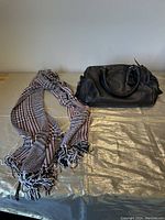 Black leather purse sitting next to a black and taupe plaid scarf on a beige surface.