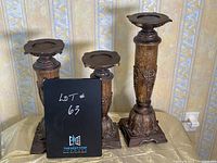 Three dark brown pillar candle holders with ornate designs and square bases displayed together on a fabric-covered table in front of patterned wallpaper.