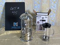 Both coffee presses on table, large with stainless steel parts and small with box in background