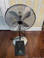 Front view of silver-colored extendible floor fan placed on wooden floor, showing four metal blades enclosed in a circular metal cage, round base, and adjustable pole height.