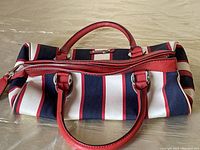 Side view of the rectangular handbag with red, white, and blue vertical stripes, red leather handles and trim showing minor wear.