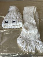 Photo showing white knit hat with faux fur pom pom and black embroidered eyelashes on cuff, next to matching white knit scarf with fringe.
