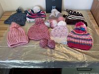 Full view of the assortment of 9 knitted touques laid flat on a table, showing different colors, pom poms, and knit styles.