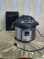 Instant Pot pressure cooker plugged in, showing the control panel and outer body with original box behind.