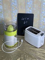 Photo showing citrus juicer and Rival toaster on wooden surface against patterned wallpaper, with Lot #69 notebook in the background.