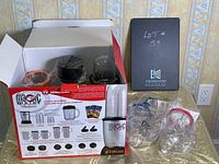 Photo showing open box with Magic Bullet components inside including power base, several cups and blades wrapped in plastic, and original packaging.