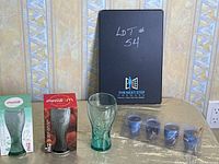 Photo showing three Coca-Cola glasses, two in their original boxes (one green, one red) and one loose glass, next to four blue Pepsi candles in plastic packaging.