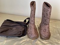 Pair of grey floral boots with brown plush lining standing upright beside a brown nylon bag with shoulder strap.