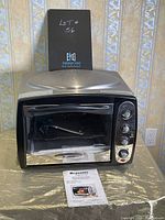Front view of Bravetti Jet Portable Convection Oven with instruction manual placed beneath it, showing control knobs on the right and glass door.