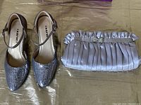 Pair of silver sling back shoes visible from top with sequined toes and ankle straps, next to silver pleated cloth evening bag with rhinestone ornament