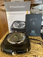 Toastmaster electric single burner hot plate shown with original box and a black sign showing lot number 3 placed beside it. The burner unit is black with a metal coil and a control knob on front.