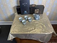 Overview of the entire tea/sake set including teapot, four cups, and original packaging boxes on a table with a gold cloth.