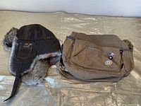 Pair of items on metallic surface: Windriver furry winter hat and brown nylon bag.